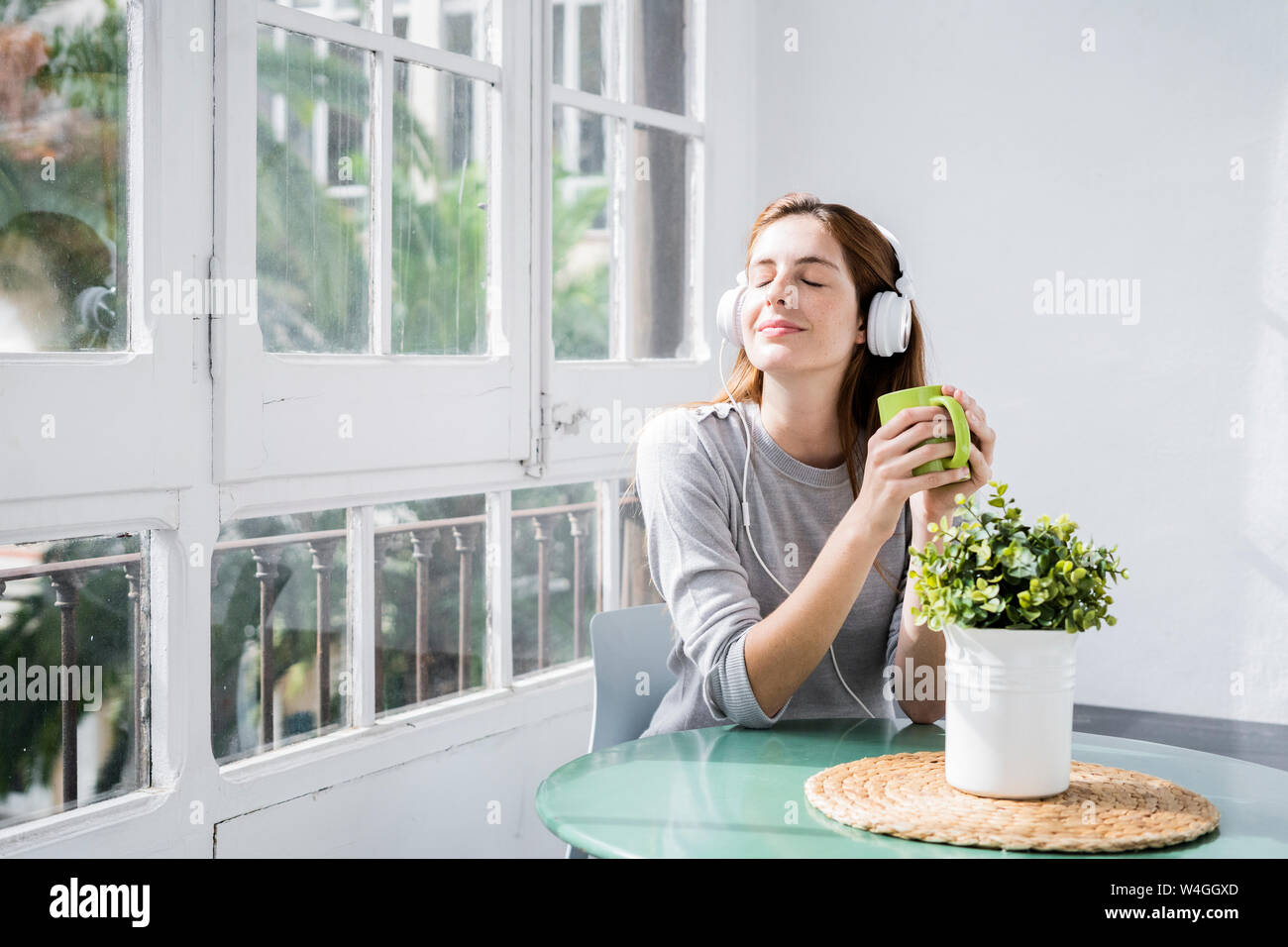 Young woman stting at the window at home with coffee cup and headphones Stock Photo