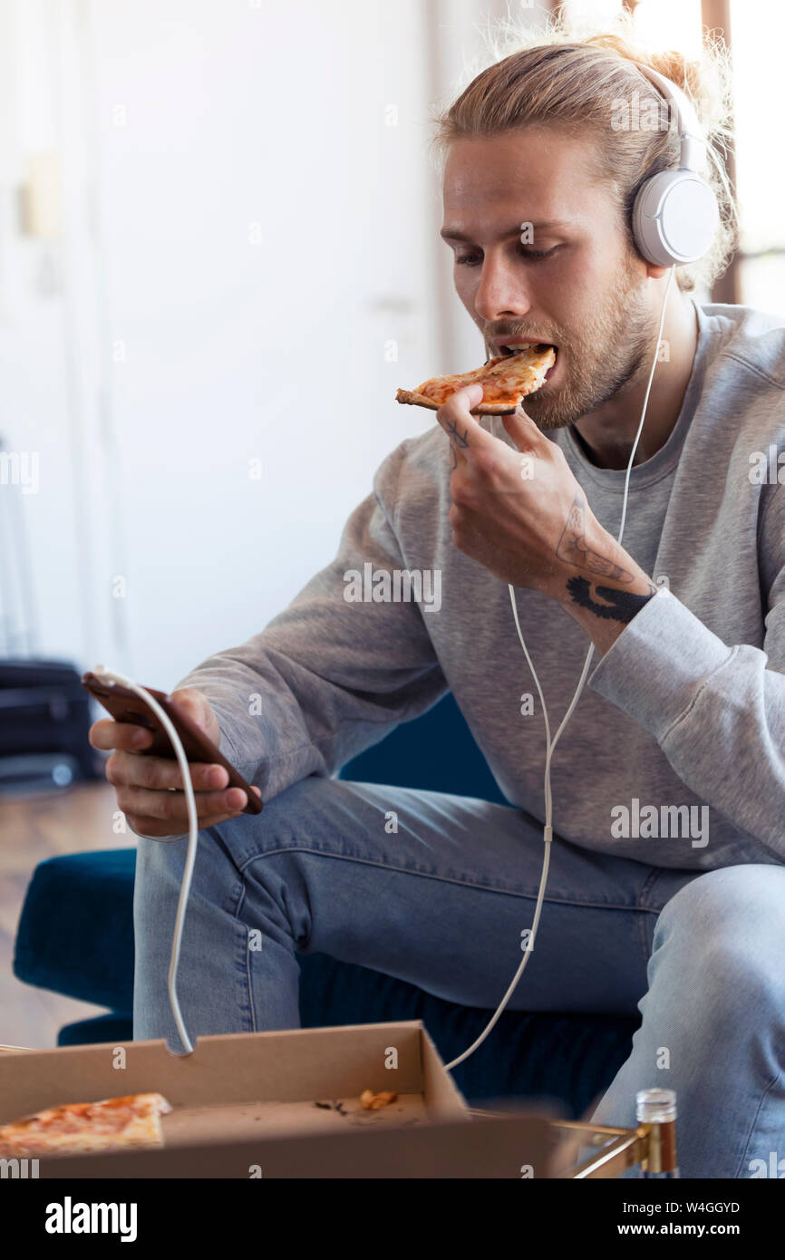 Young man on couch with cell phone and headphones eating pizza Stock Photo