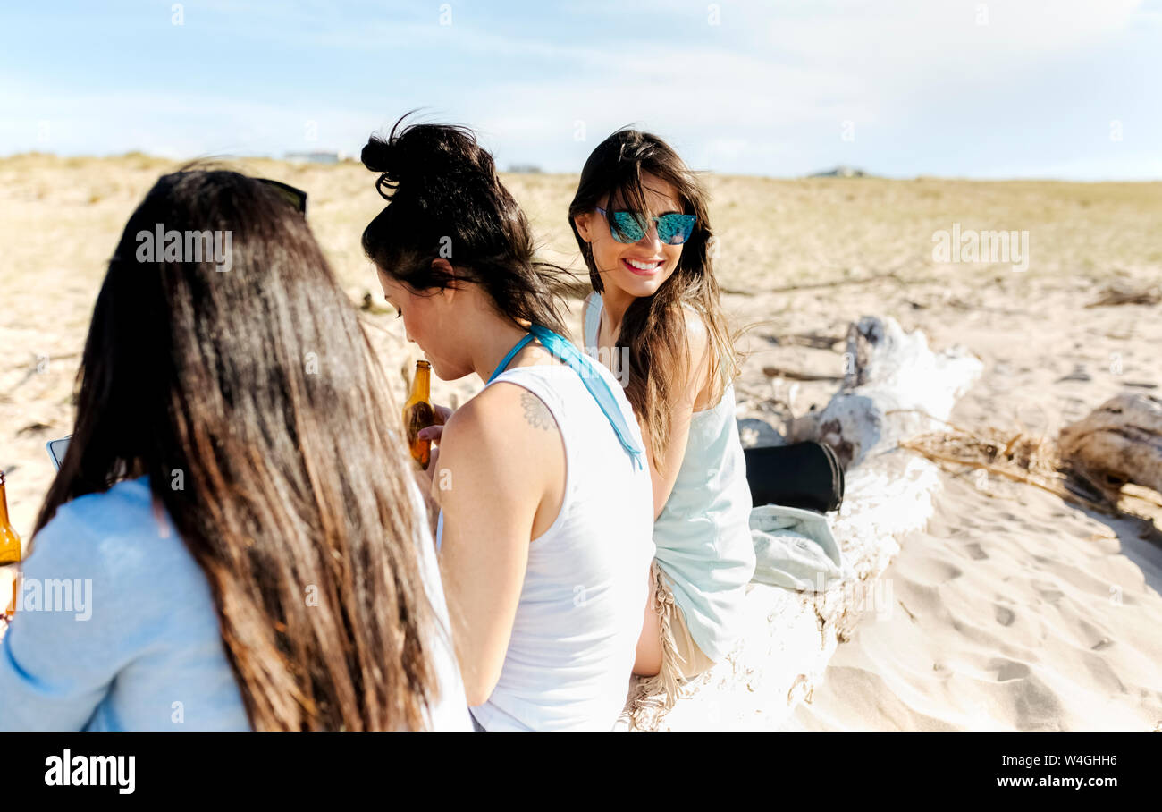 Happy female friends sitting on log on the beach Stock Photo
