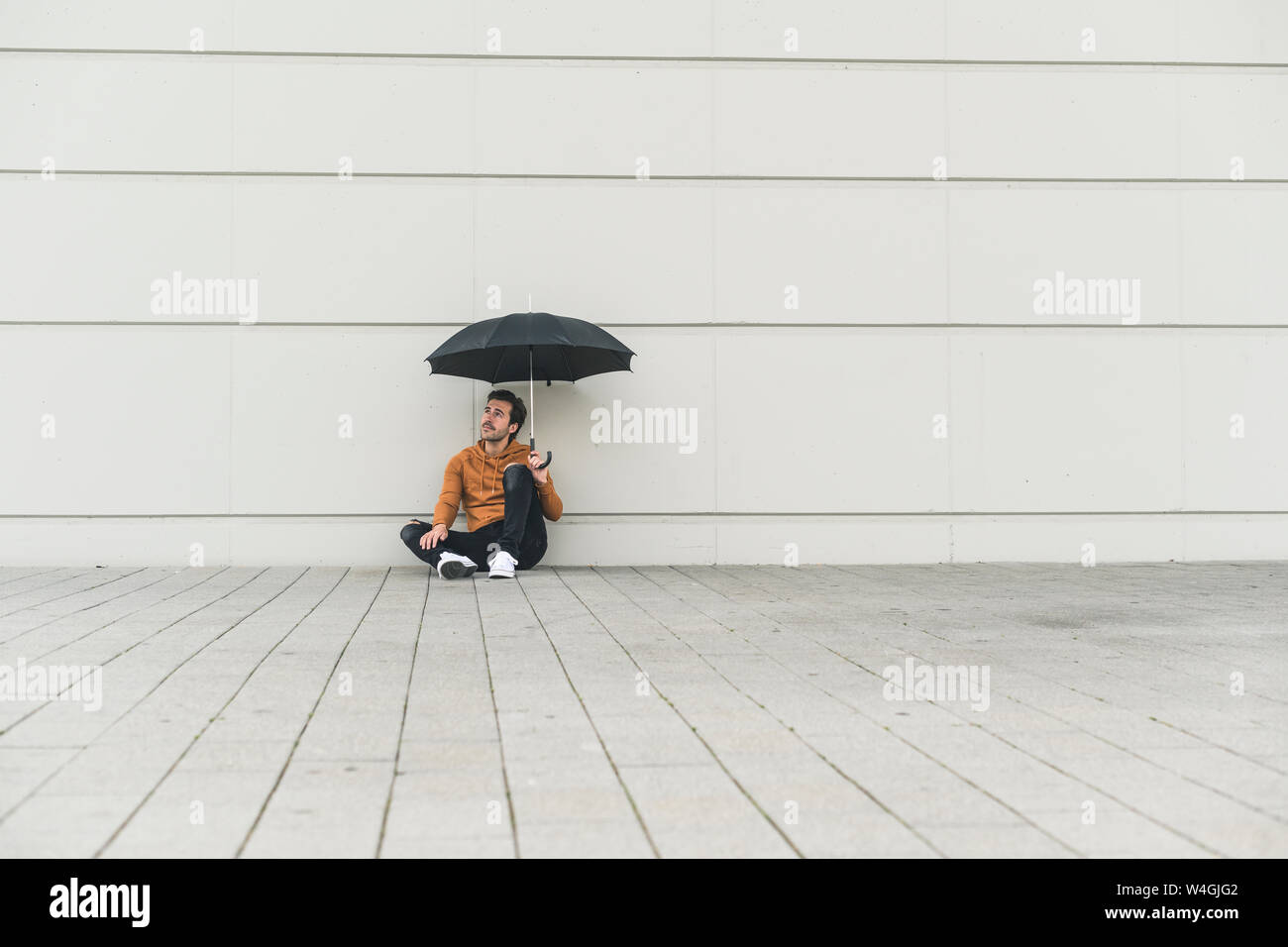 Young man with umbrella, sitting on ground, looking uncertain Stock Photo