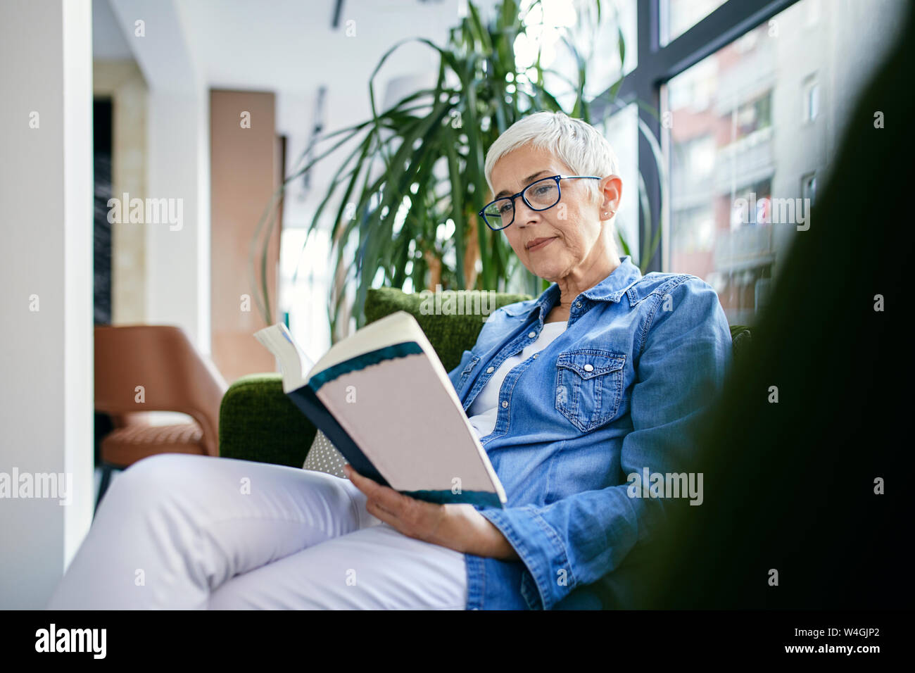 Senior woman sitting on couch, reading a book Stock Photo - Alamy