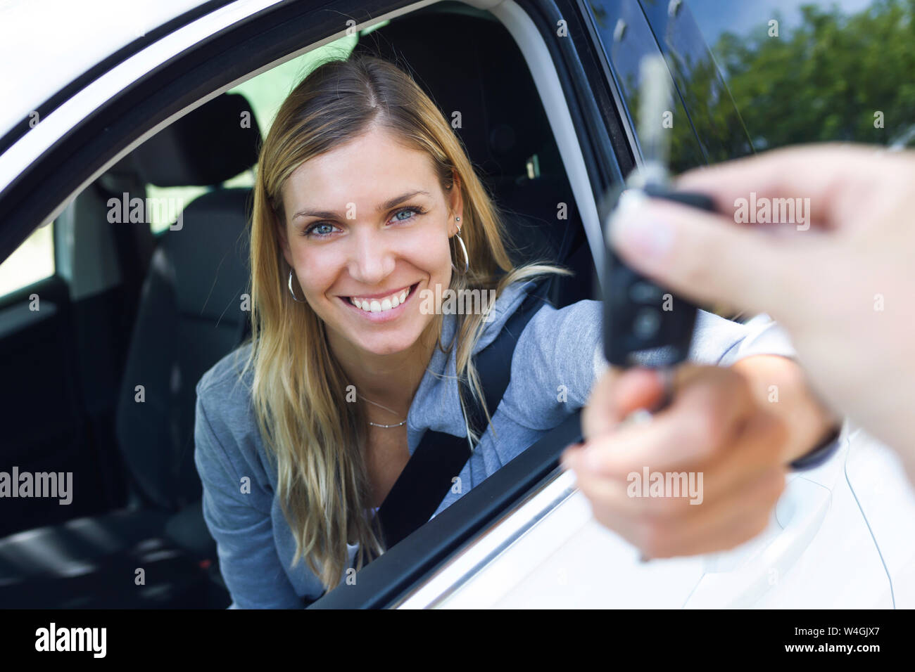 Portrait of smiling young woman looking at camera while holding car keys and give it to someone through the window Stock Photo