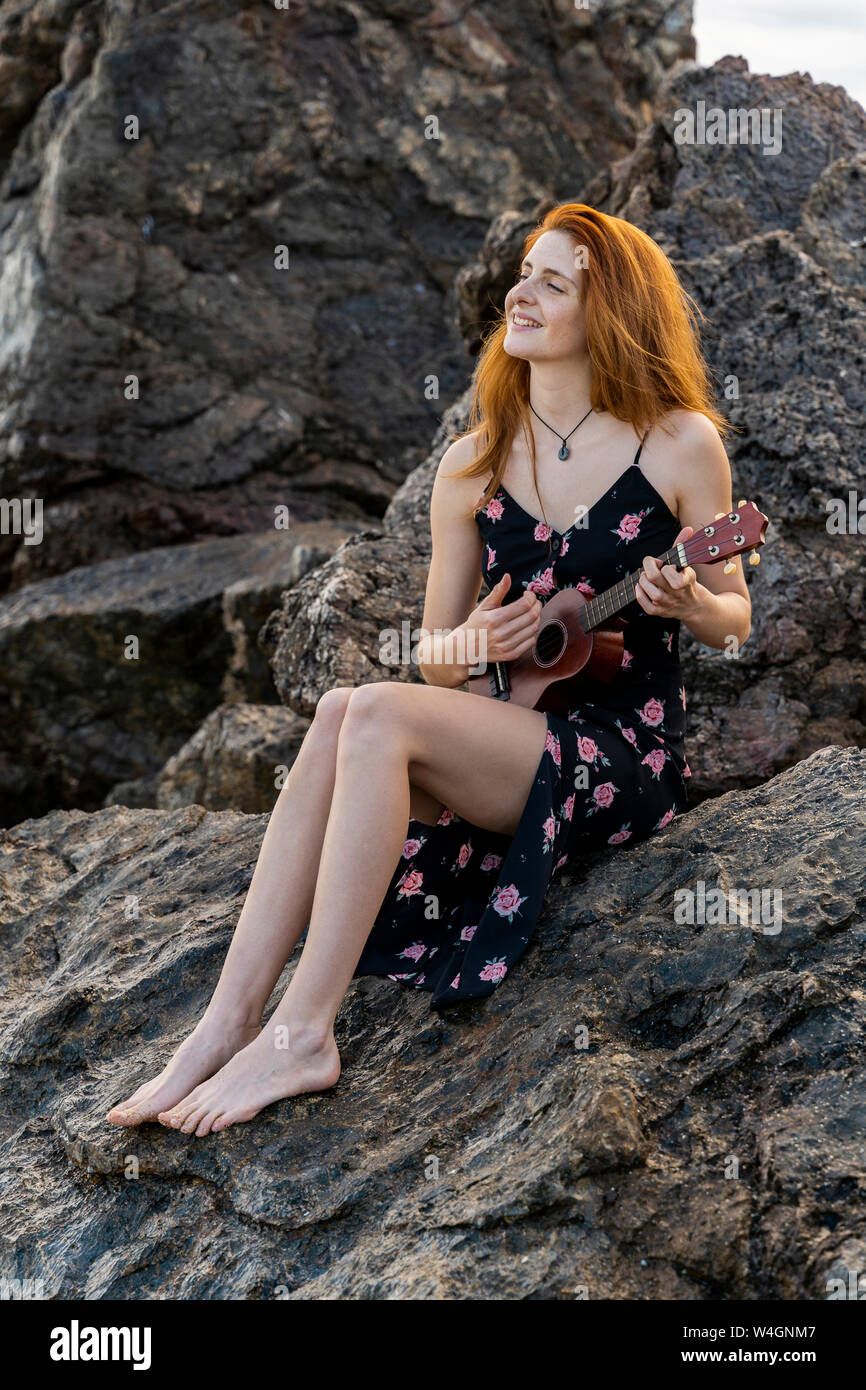 Smiling redheaded young woman sitting on rock playing ukulele Stock Photo