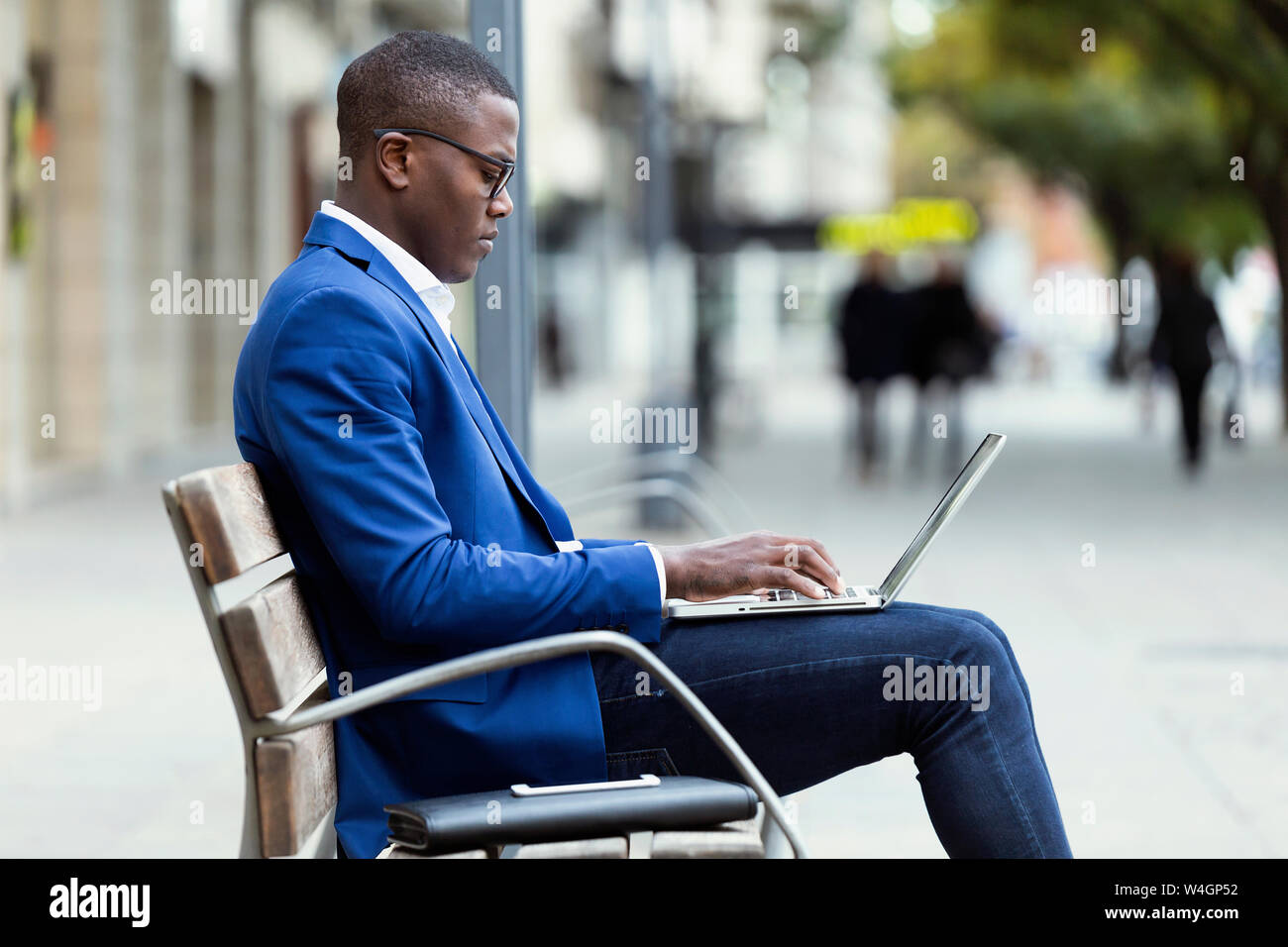 Young businessman wearing blue suit jacket sitting on bench and using laptop Stock Photo