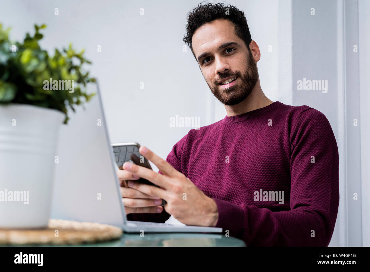 Portrait of young man stting at table at home using laptop and cell phone Stock Photo