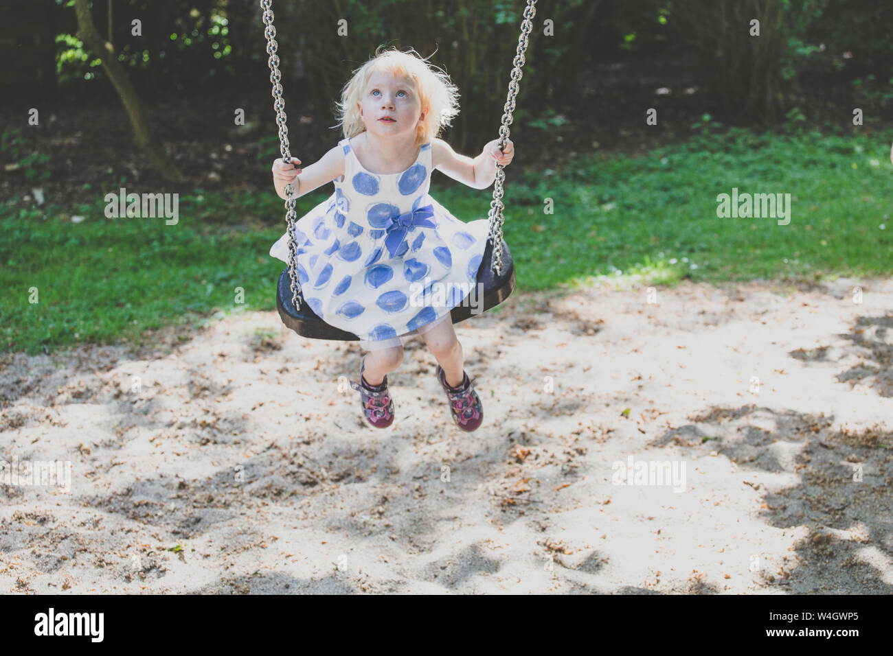 Portrait of blond little girl wearing summer dress sitting on a swing looking up Stock Photo