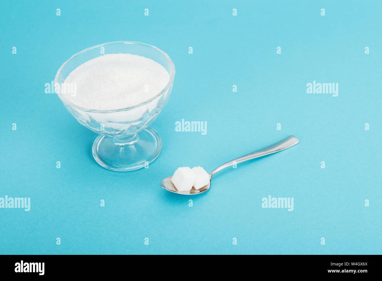 glass bowl with granulated sugar and teaspoon with sugar cubes on blue