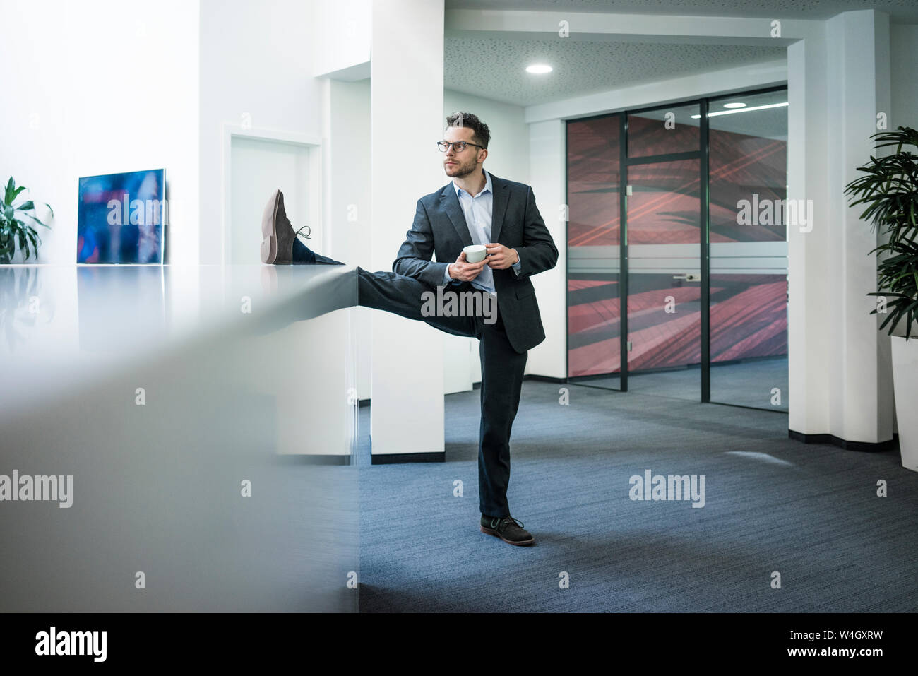 Businessman with cup of coffee standing on one leg in office Stock Photo