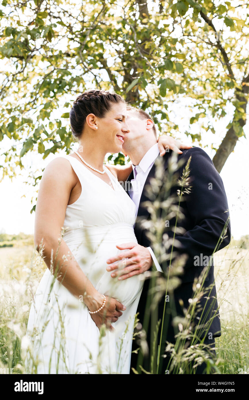 Pregnant bride with her husband holding baby belly on a meadow Stock Photo