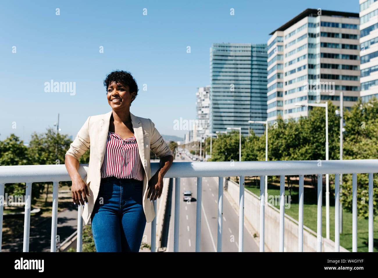 Smiling businesswoman leaning on a bridge railing, office buildings in the background Stock Photo