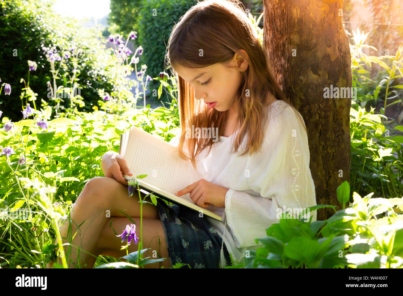 Little girl leaning against tree trunk reading a book Stock Photo