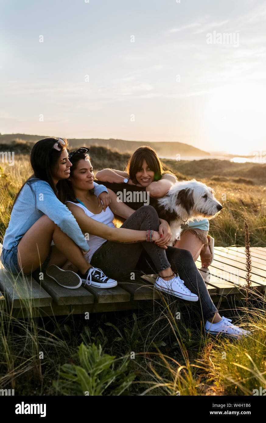 Three happy women with dog sitting on boardwalk in dunes at sunset Stock Photo