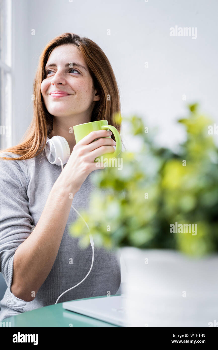 Smiling young woman stting at table at home with coffee cup and laptop Stock Photo