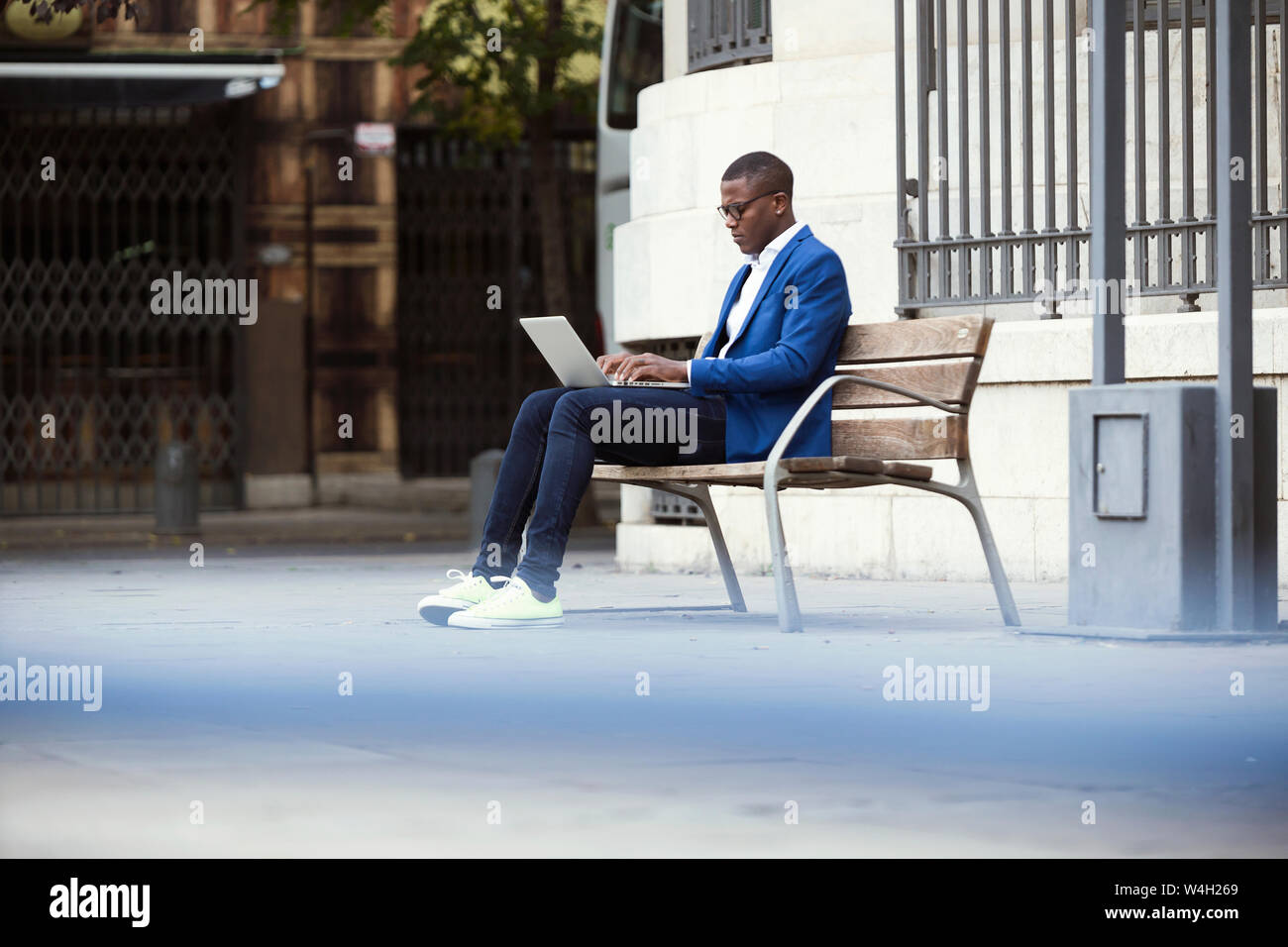 Young businessman wearing blue suit jacket sitting on bench and using laptop Stock Photo