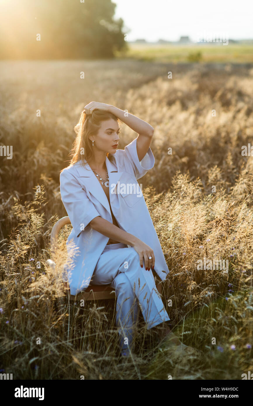 Fashion photo of attractive blonde woman in blue fashion suit sitting on chair in wheat field. Stock Photo