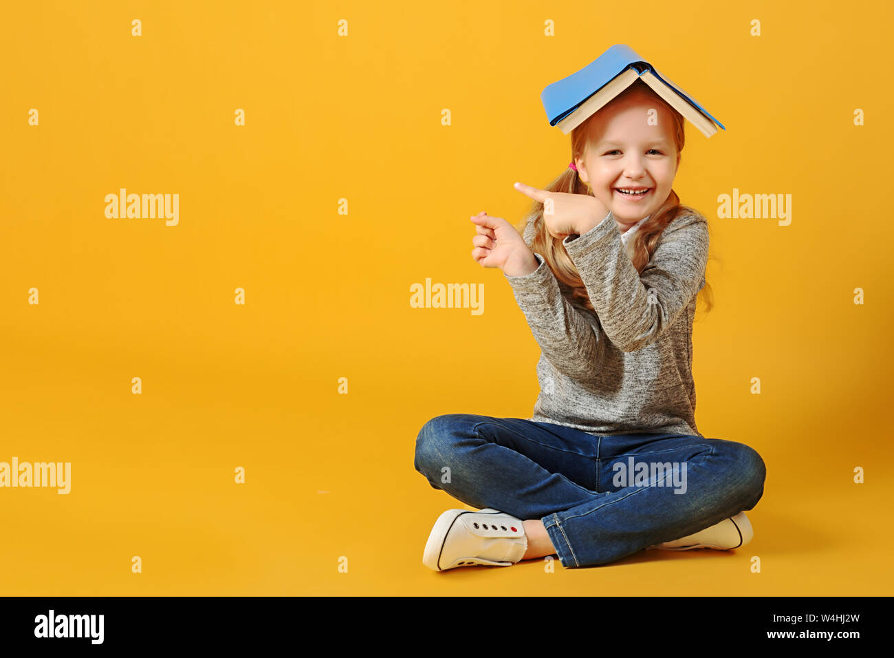 Cheerful attractive little student girl is sitting on the floor with a book on her head and pointing to the side. The concept of education and school. Stock Photo