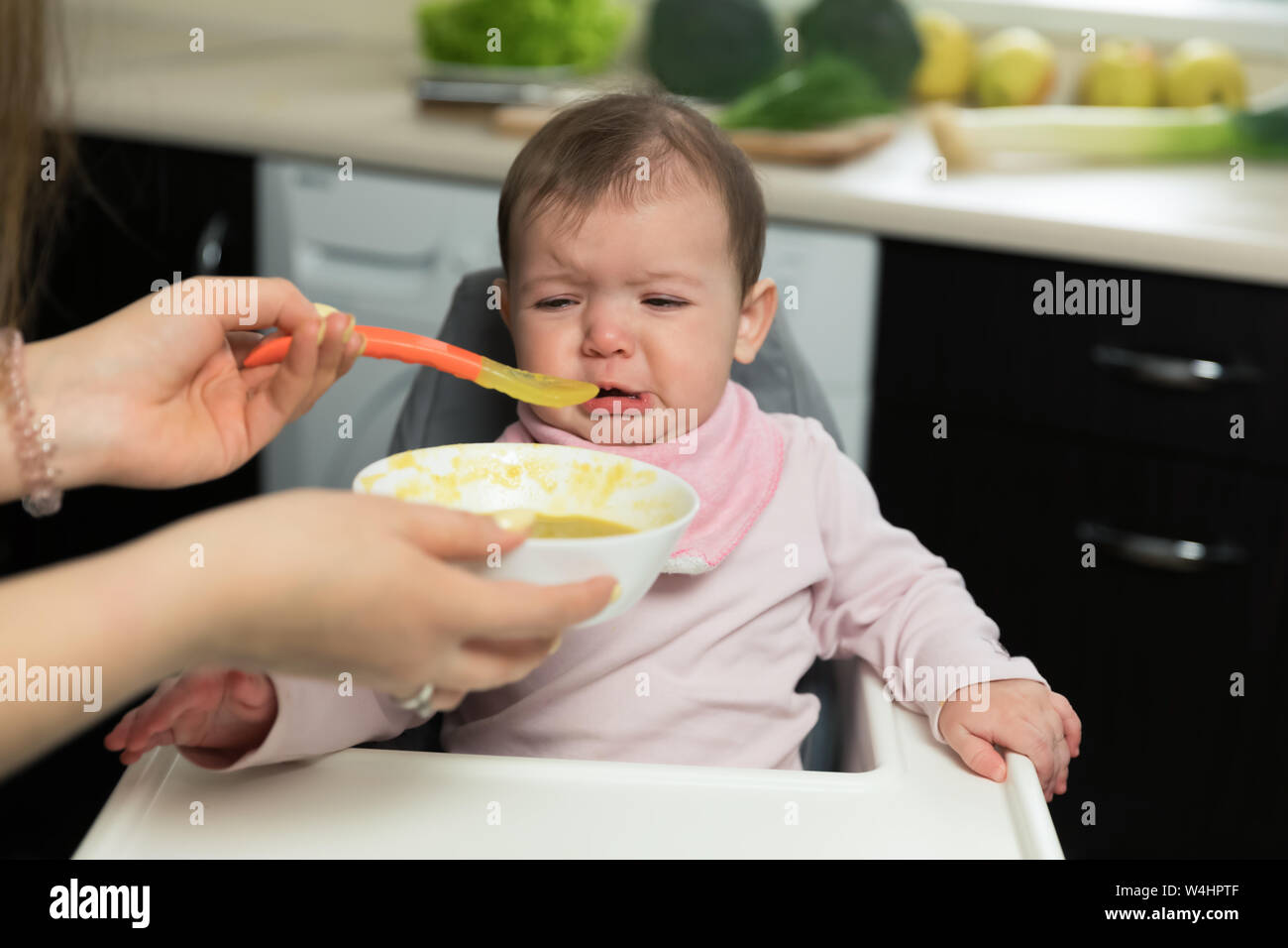 Mom feeds a baby with a spoon. The baby is crying while sitting at