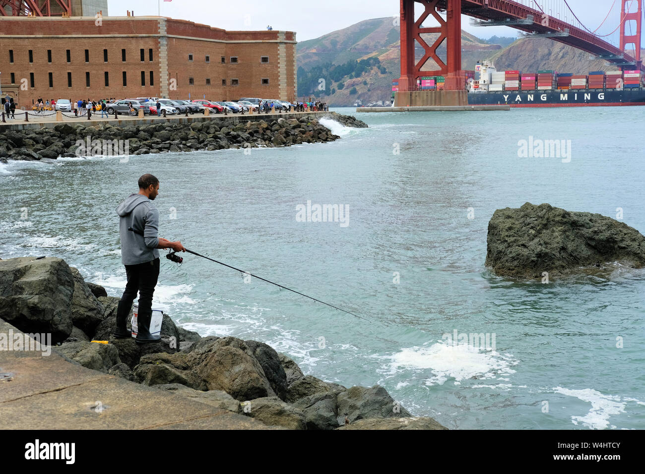 Young African American male fishing while a Yang Ming marine transport vessel sails under the Golden Gate Bridge in San Francisco, California. Stock Photo