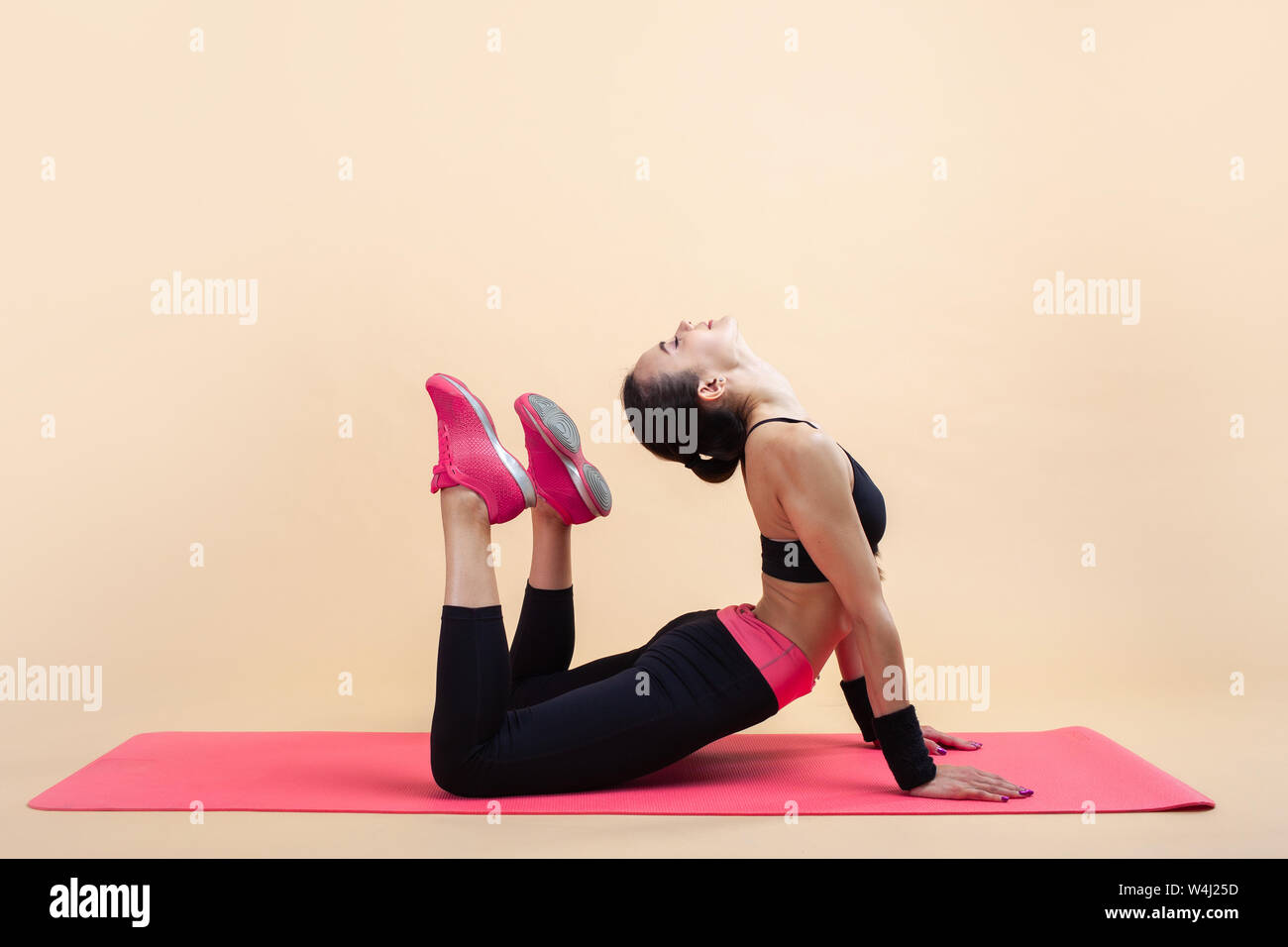 A young attractive girl with a ponytail, brunette, is lies in a cobra pose with knees bent, doing stretching on a pink yoga mat, on a pink background. Stock Photo