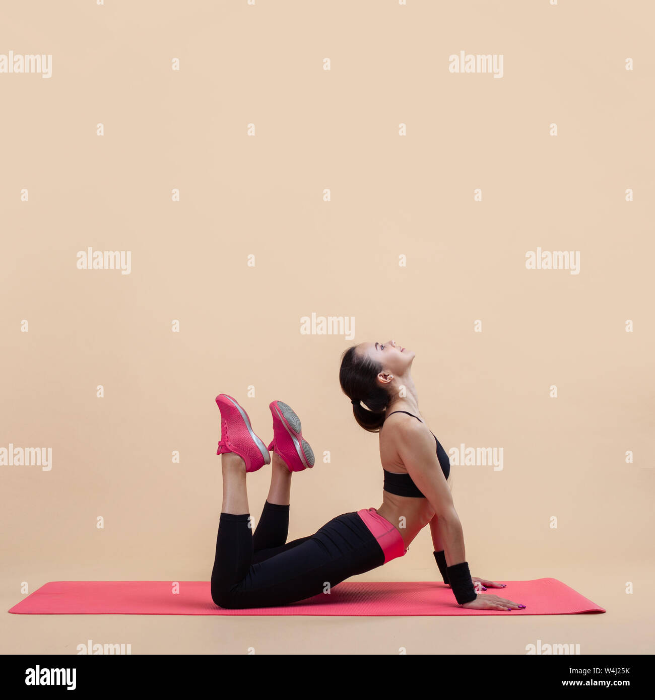 A young attractive girl with a ponytail, a brunette, is lies in a cobra pose with knees bent, doing stretching on a pink yoga mat, on a pink backgroun Stock Photo