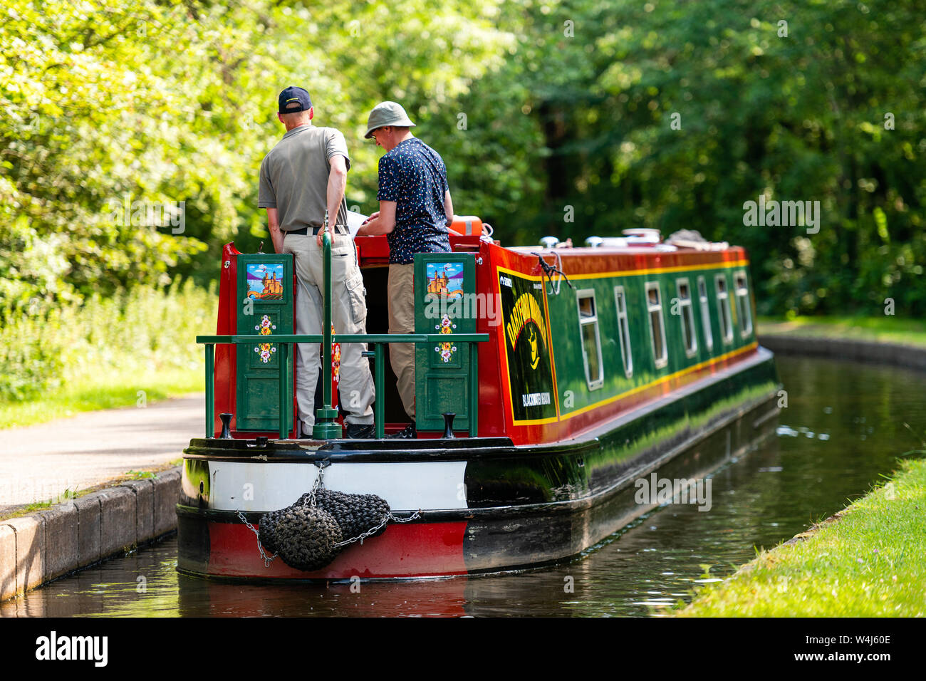 Rear view of two men on the stern of a narrow boat navigating along a canal on a sunny summer day, UK. Stock Photo