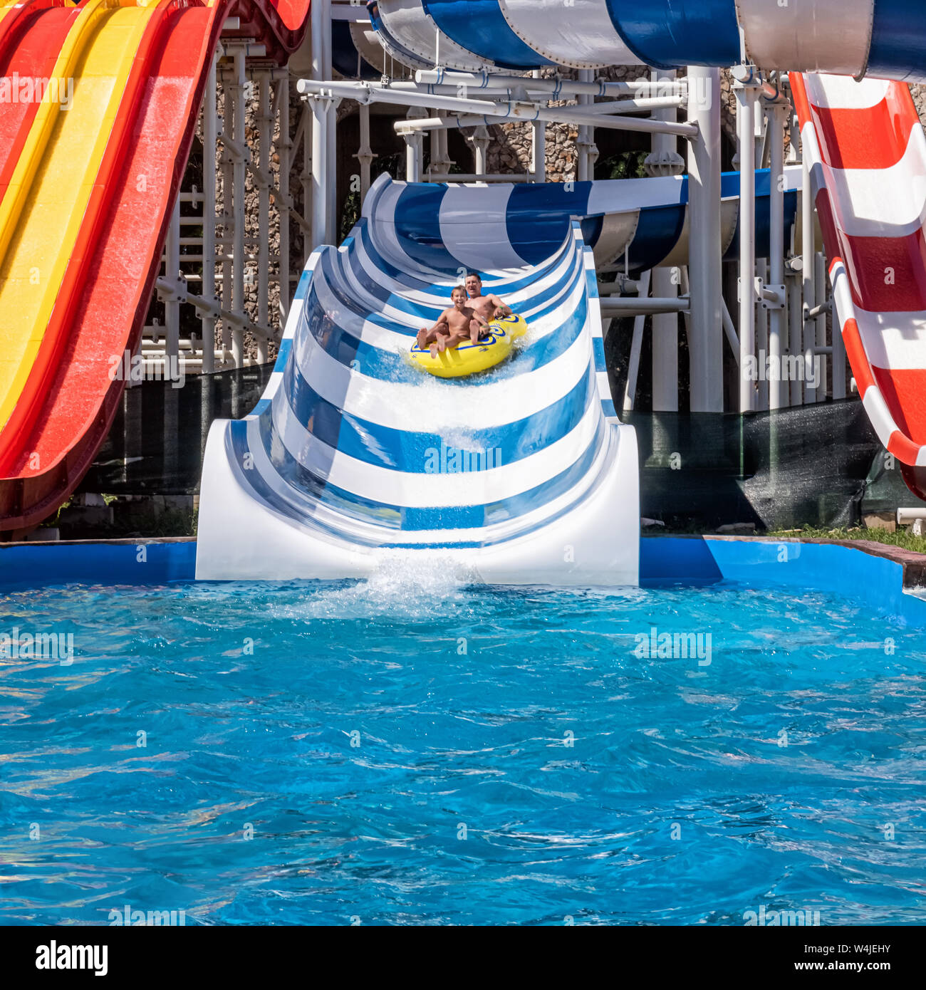 Happy laughing father and son at amusement park. Adult and teenager boy are enjoying weekend together, riding down the water slide in yellow inflatabl Stock Photo