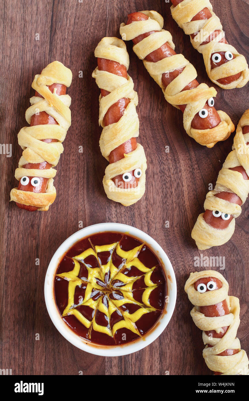 Fun food for kids. Mummy hot dogs lying on a rustic table with a bowl of ketchup and mustard dip, with spider web design. Top view, flay lay position. Stock Photo