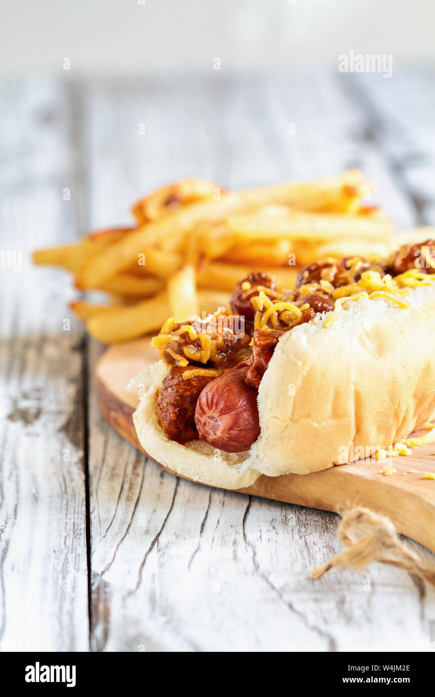 Hot dog with chilli, cheddar cheese and mustard. Selective focus with blurred french fries in the background. Stock Photo