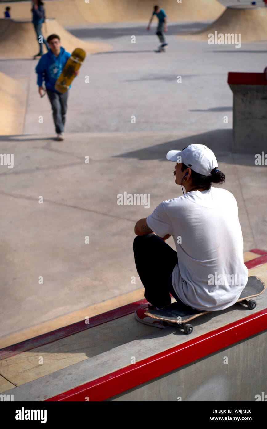 A young man sitting alone, watching kids skate from a distance at the