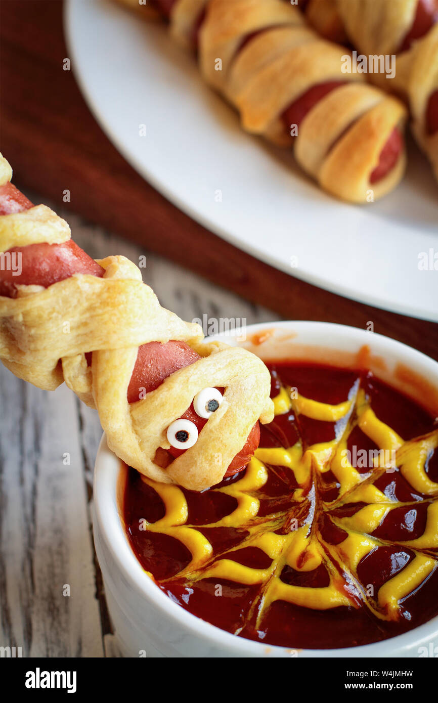 Fun food for kids. Mummy hot dogs held upside down over a bowl of ketchup and mustard dip, with spider web design. Stock Photo