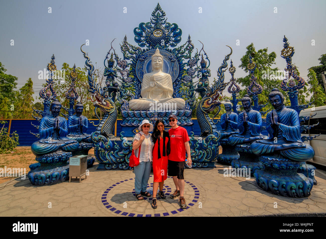 Tourists pose in front of a seated, white statue of Buddha sitting on his throne surrounded by statues of priests at the Blue Temple, Wat Rong Seua Te Stock Photo