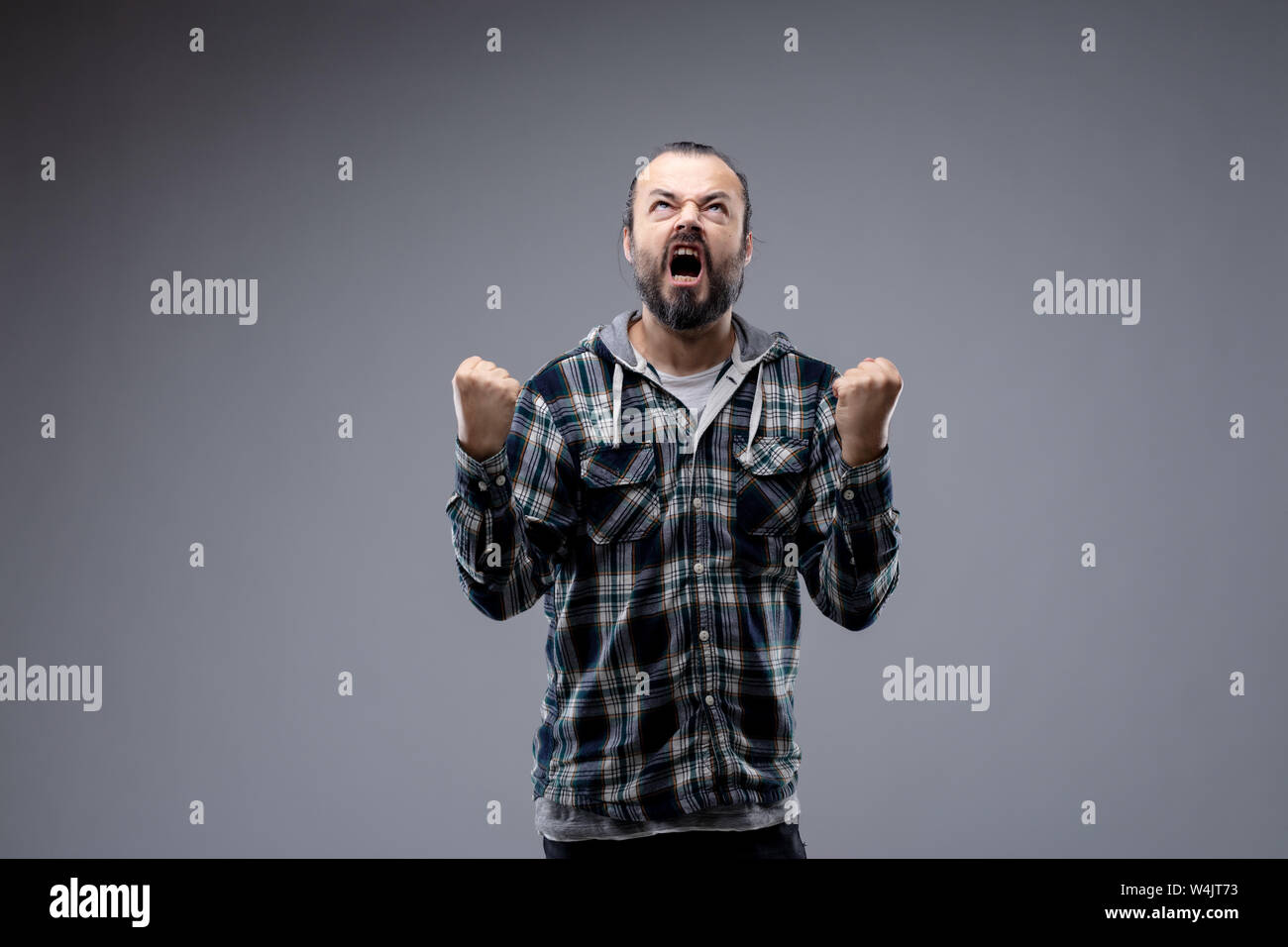 Angry man throwing a temper tantrum clenching his fists and yelling into the air with an expression of fury isolated on grey with copy space Stock Photo