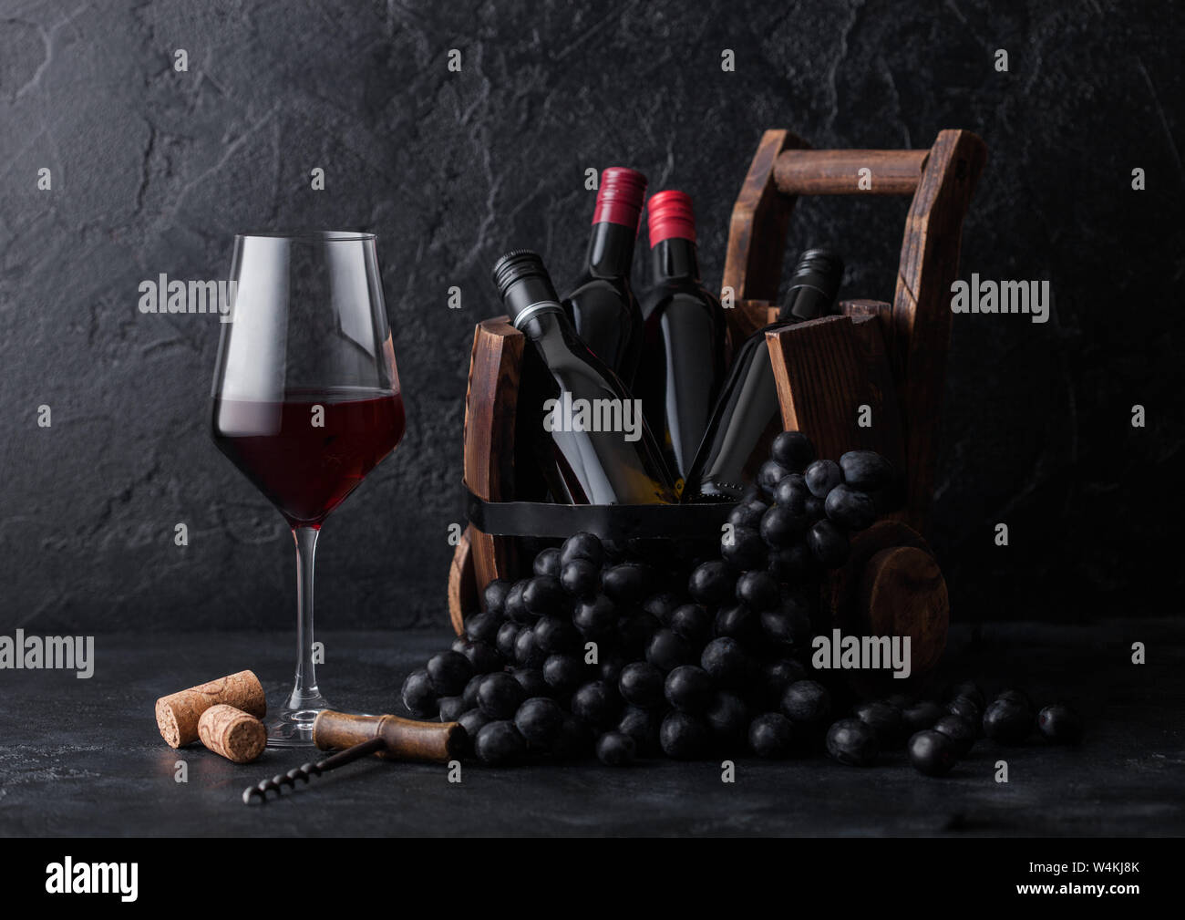 Elegant glass of red wine with dark grapes and mini bottles of wine inside vintage wooden barrel on black stone background. Stock Photo