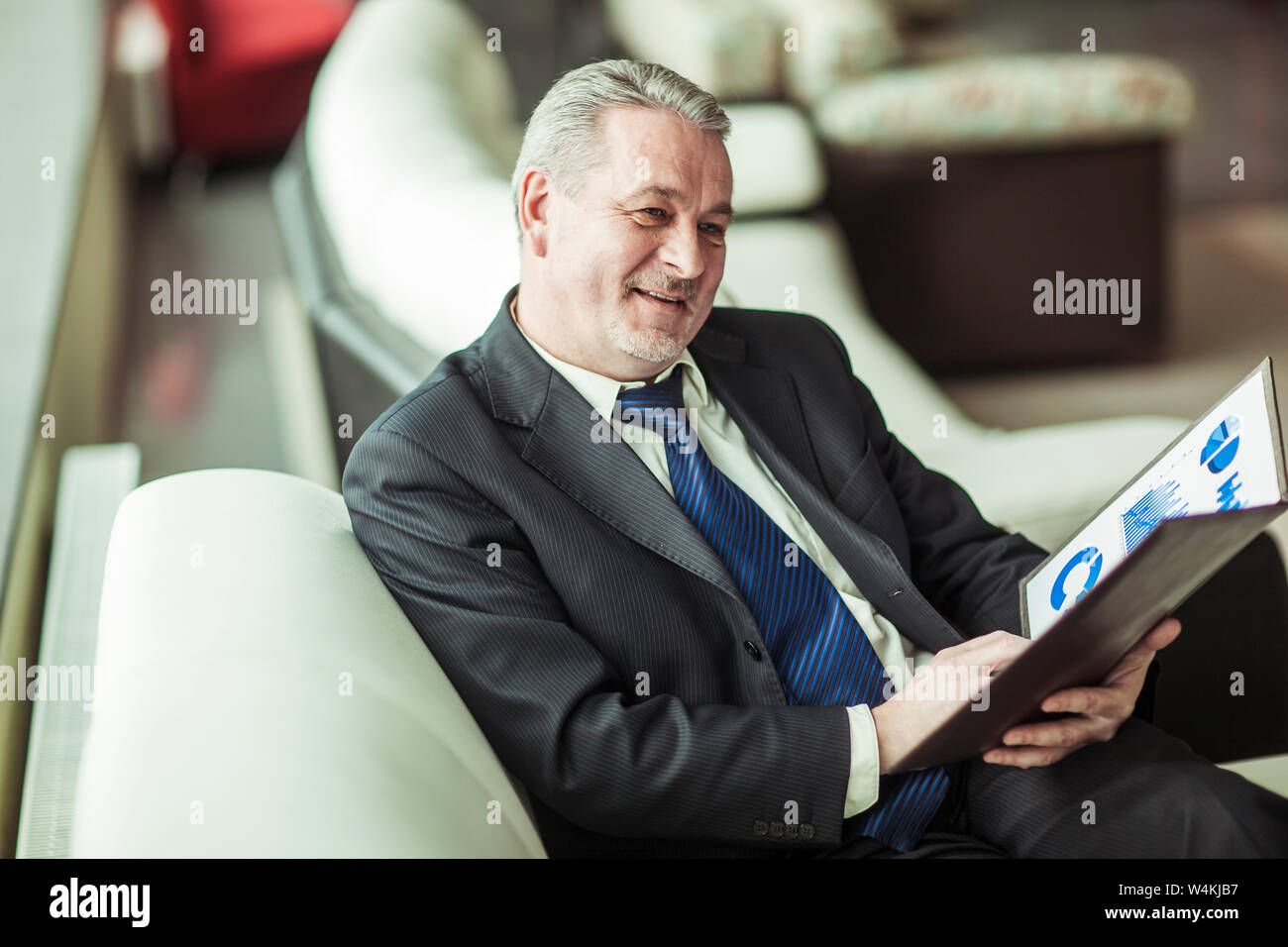 foreground is an experienced financier with financial charts sitting in the chair in front of the office. Stock Photo