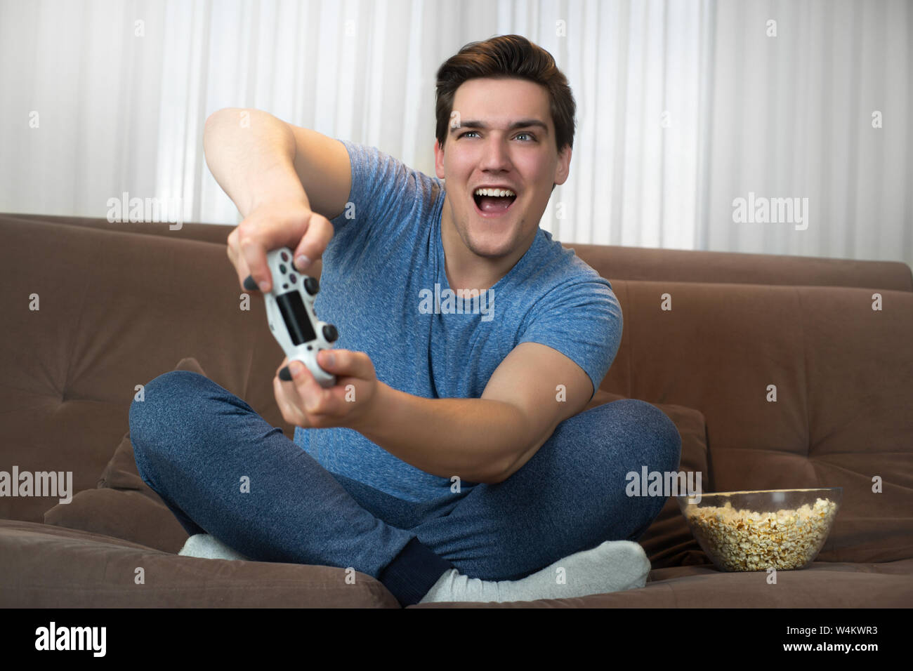 young handsome man playing video game sitting on comfy sofa holding joystick with both hands looking involved in mission Stock Photo