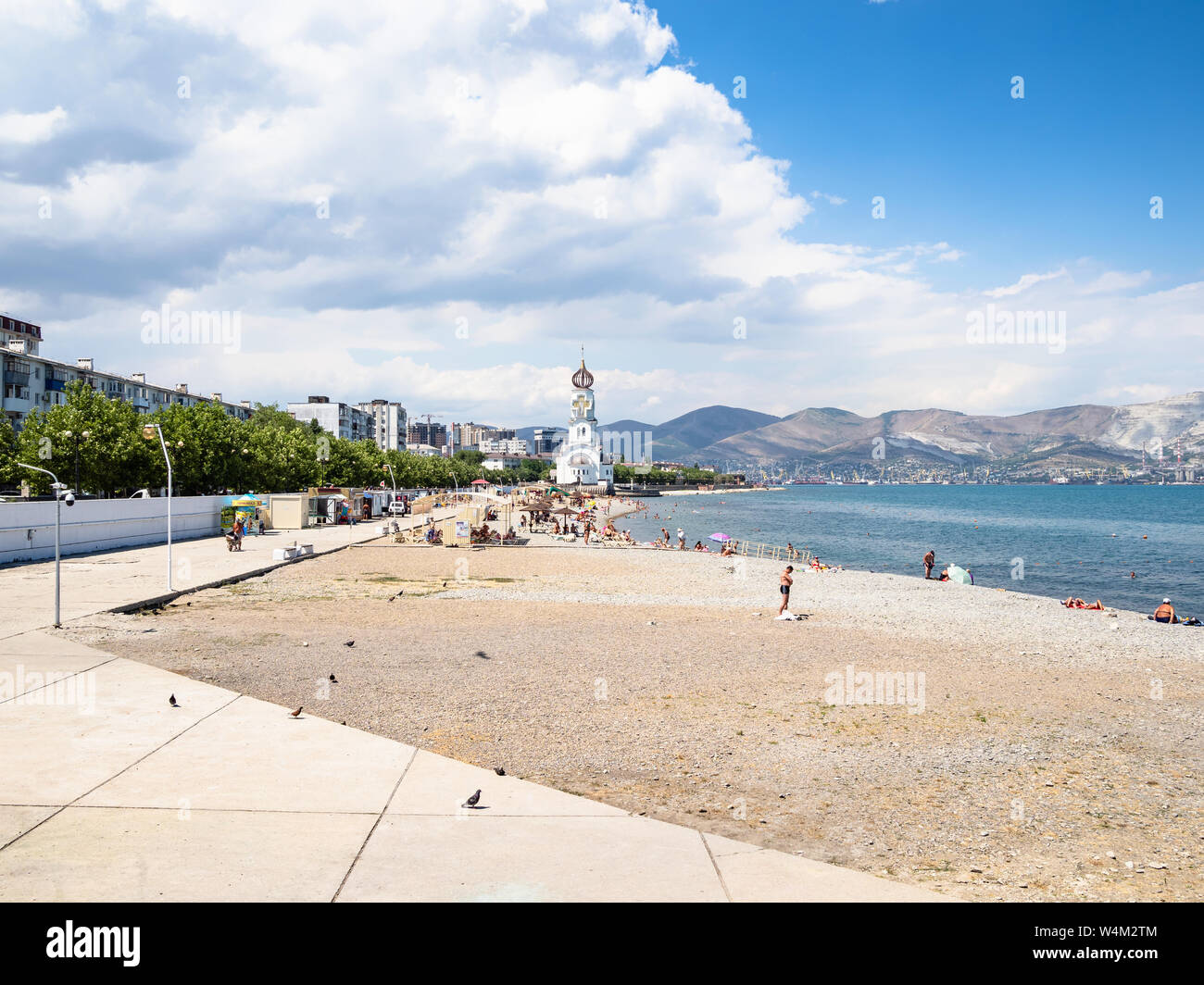 NOVOROSSIYSK, RUSSIA - JULY 7, 2019: tourists on city beach near Chapel of St Nicholas the Wonderworker and Church of St Peter and St Fevronia on Admi Stock Photo