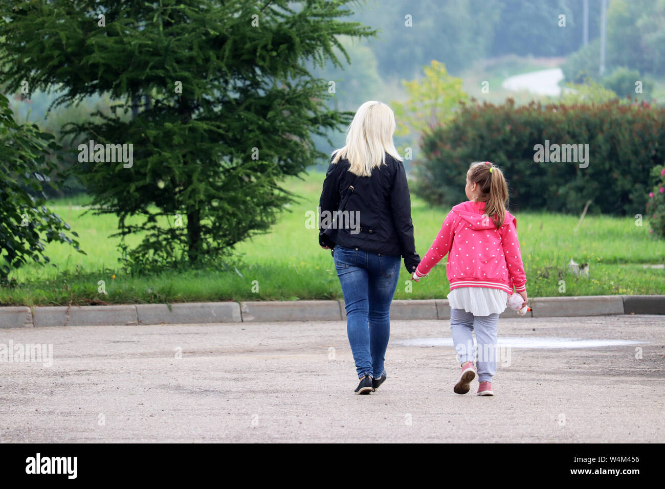 Mother and daughter are walking in a green park holding hands. Blonde woman lead little girl, concept of motherhood, single mom or babysitter Stock Photo