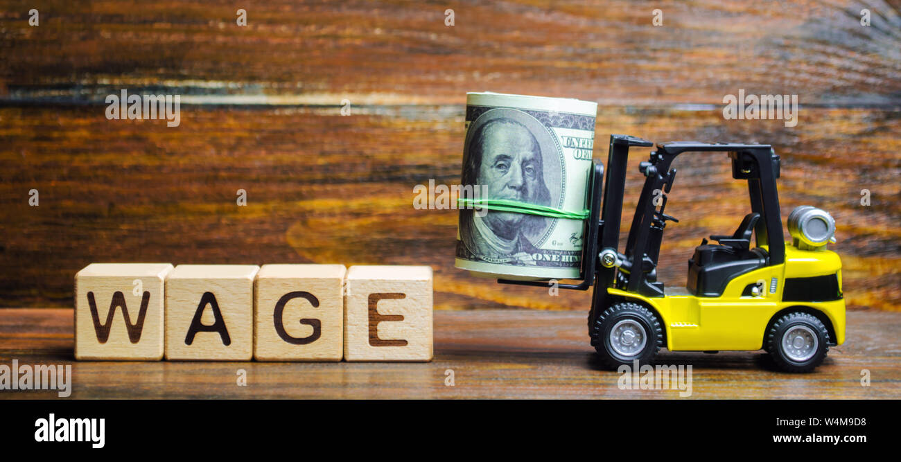 Wooden blocks with the word Wage, dollars and a forklift. Financial compensation. Salary payment to employees. Minimum and nominal wages rates. Profit Stock Photo