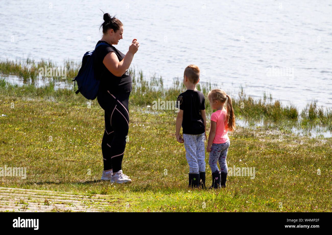 Dundee, Tayside, Scotland, UK. 24th July, 2019. UK weather: Hot sunny weather with temperatures reaching 25º Celsius.  A Polish woman taking photographs of her two children on her mobile phone beside the pond at Clatto Country Park in Dundee, UK. Credit: Dundee Photographics / Alamy Live News Stock Photo