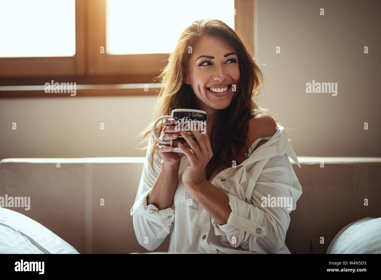 A young happy smiling woman is drinking morning coffee sitting in the room after awakening. Stock Photo