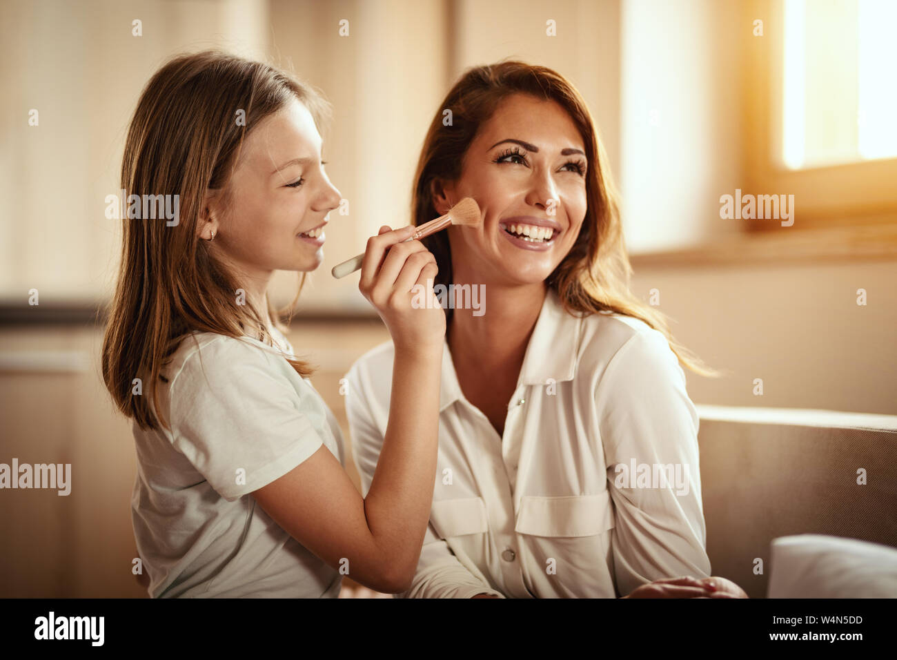 Little daughter is having fun playing beauty salon with her young mother and putting makeup on her face with big puffy brush. Stock Photo