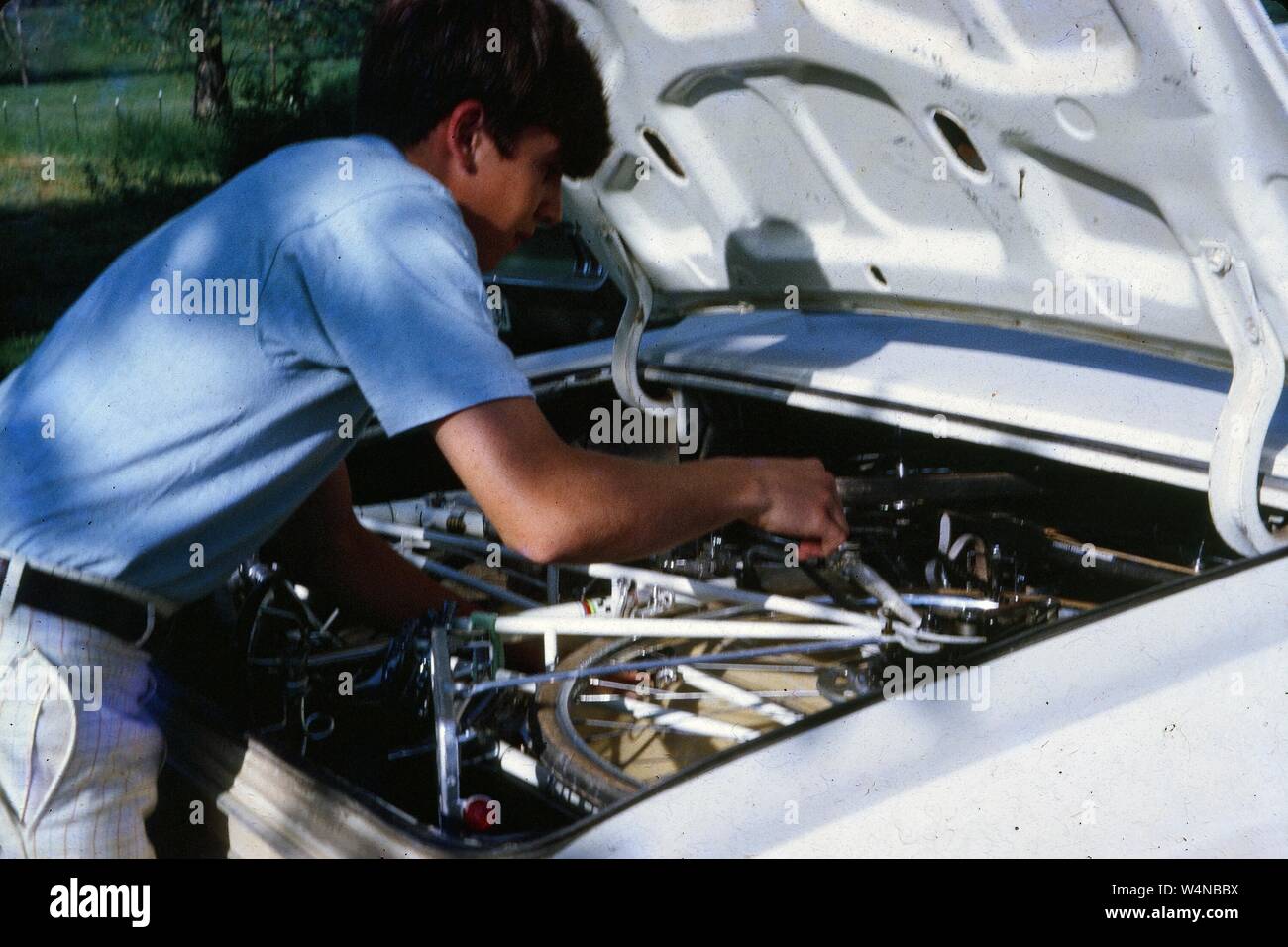 Person viewed from behind working on the engine of a car outdoors, 1970. () Stock Photo