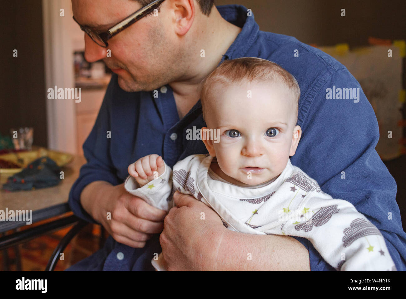 a bright-eyed baby sits in his father's lap at a dining room table Stock Photo
