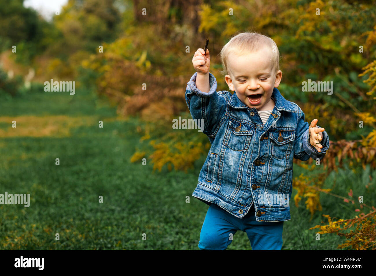 A laughing baby boy holds stick in his hand and plays outside in fall Stock Photo
