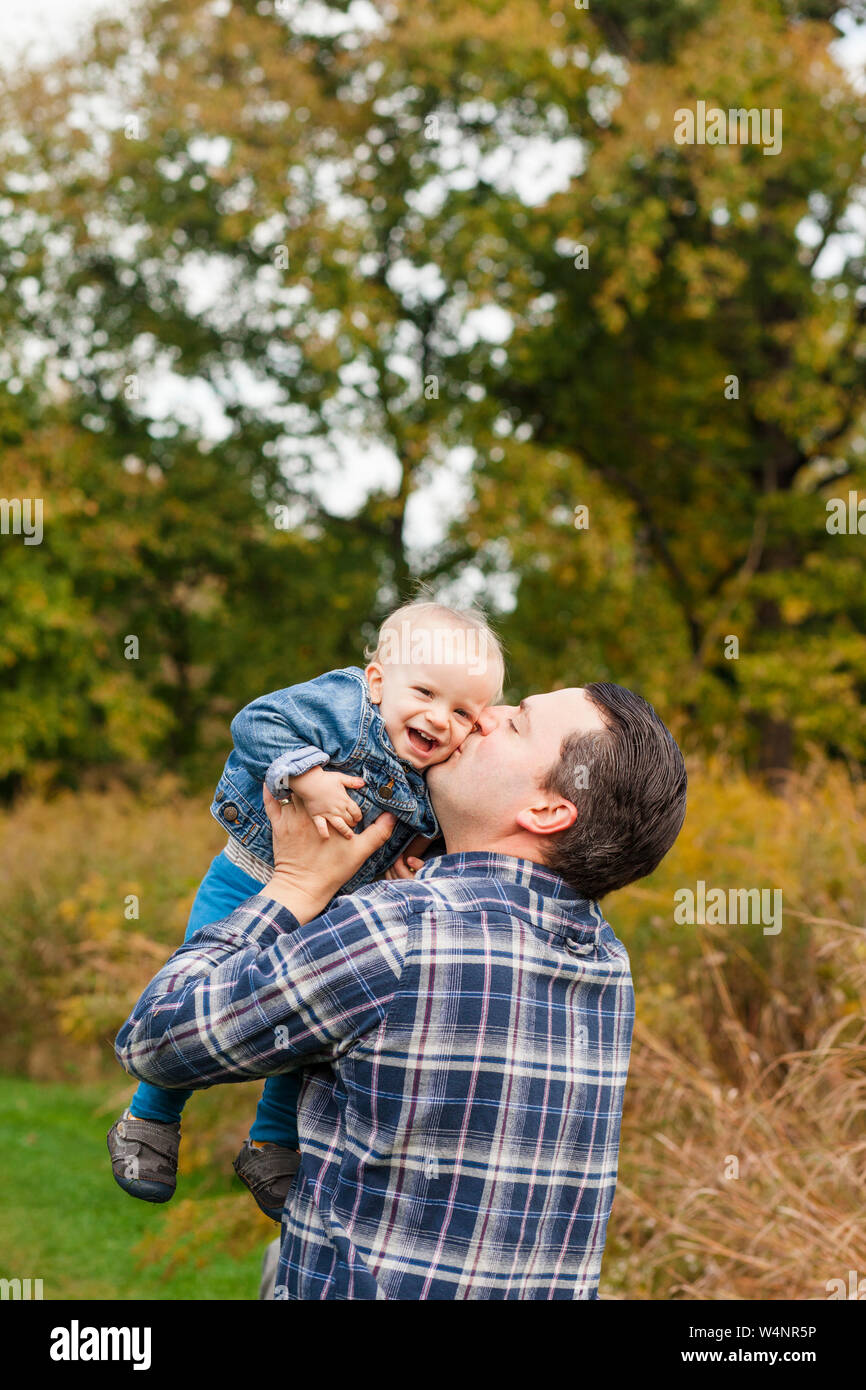 A happy father raises his laughing baby boy up to for a kiss in a park Stock Photo