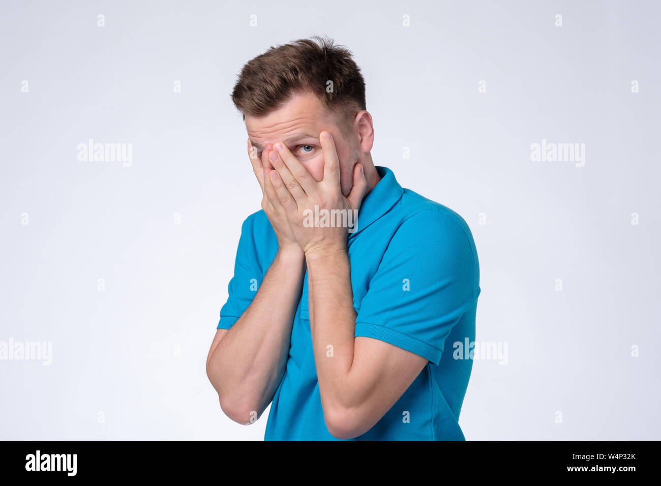 Man in blue t-shirt hiding cover face with hands, looking shy Stock Photo