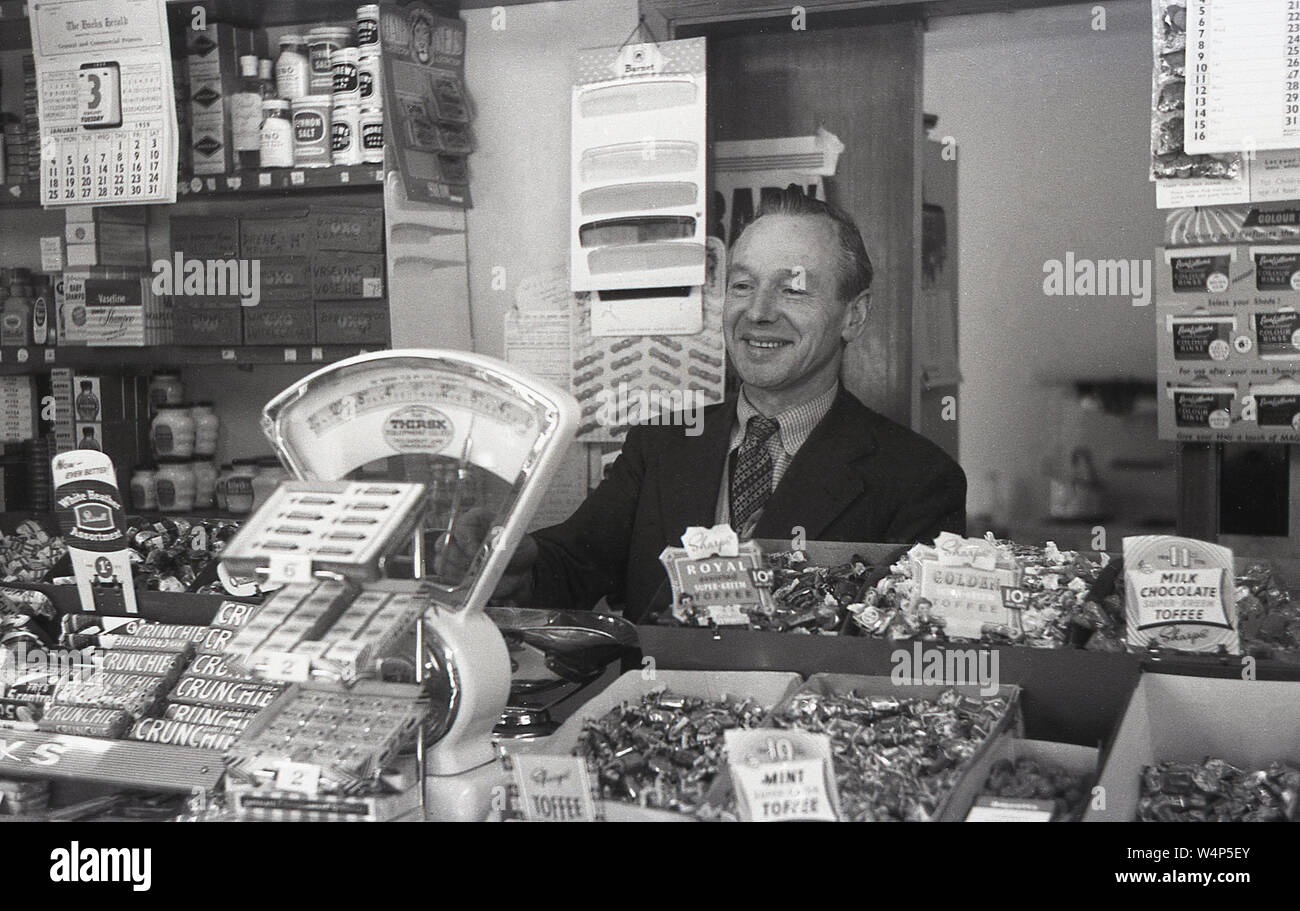 1955, historical, a newsagent owner wearing a jacket and tie behind the counter inside his shop, showing the different sweets, weighing scales and other products like combs and rubber bands tthat local shops sold in this era, England, UK. Stock Photo