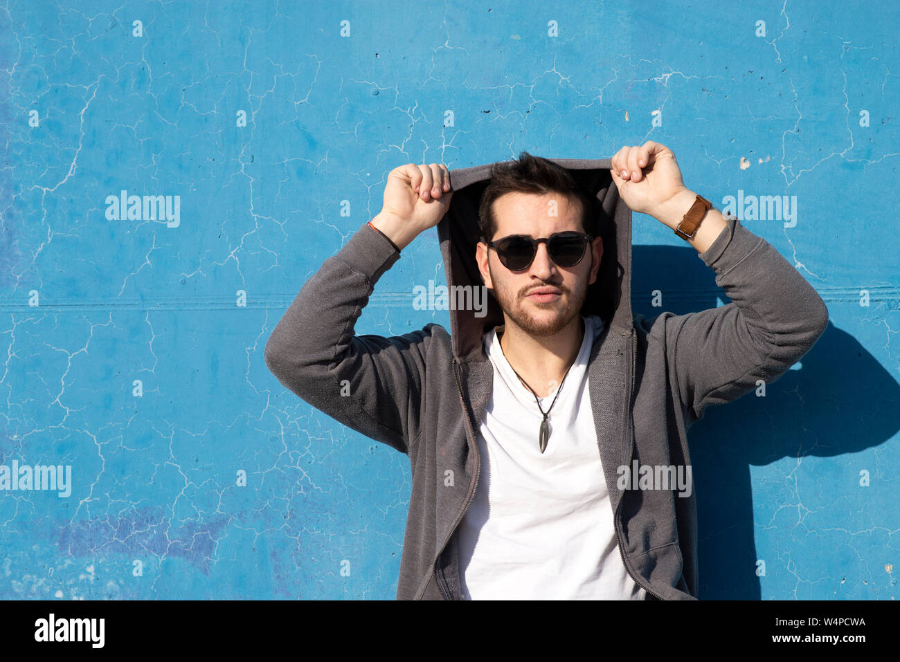 Young bearded man leaning on blue wall wearing sunglasses Stock Photo