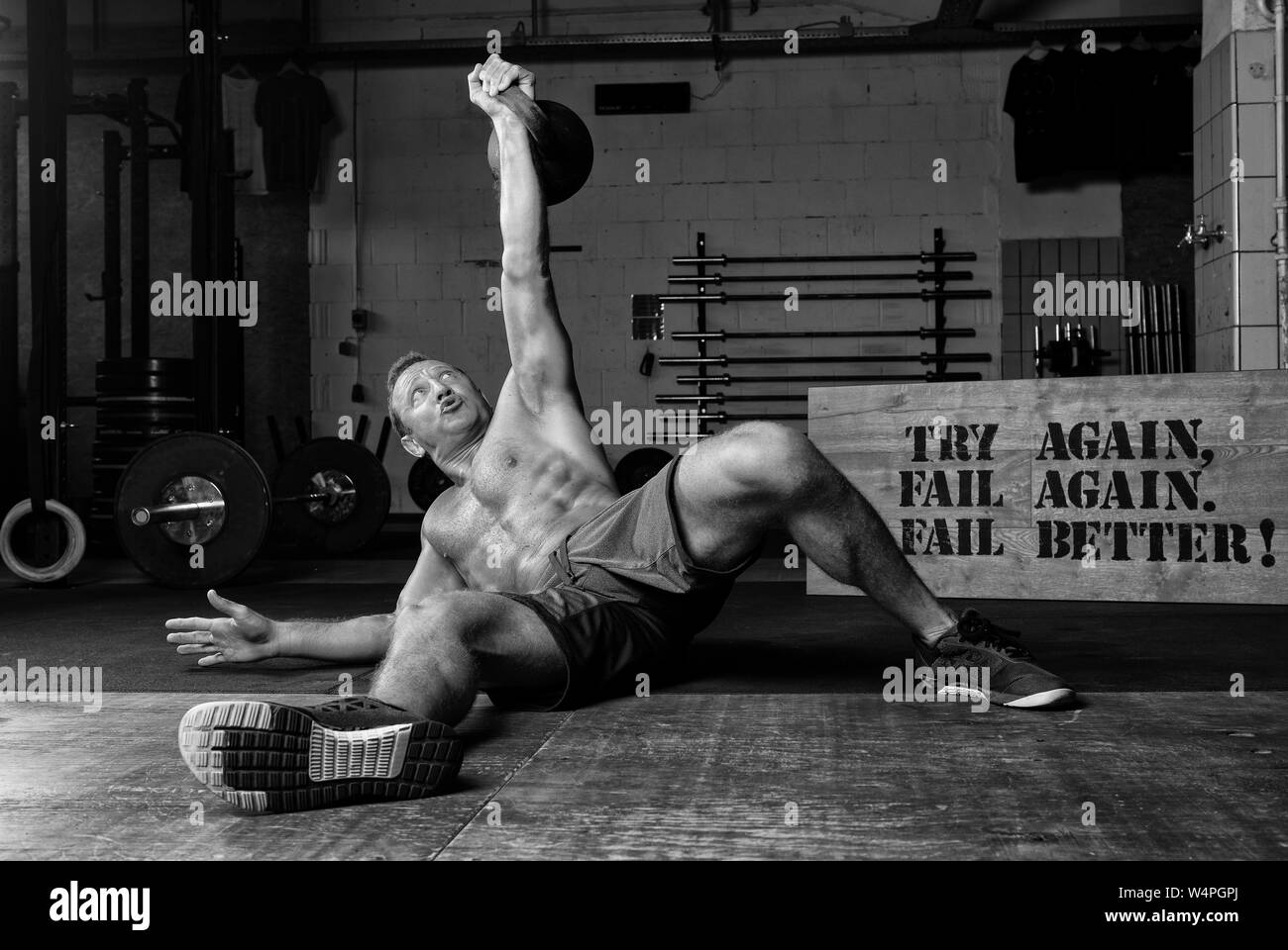 An athletic suntanned man with strong abs is doing weight lifting with a kettlebell in a gym. Exercise Turkish get up. Black and white. Stock Photo