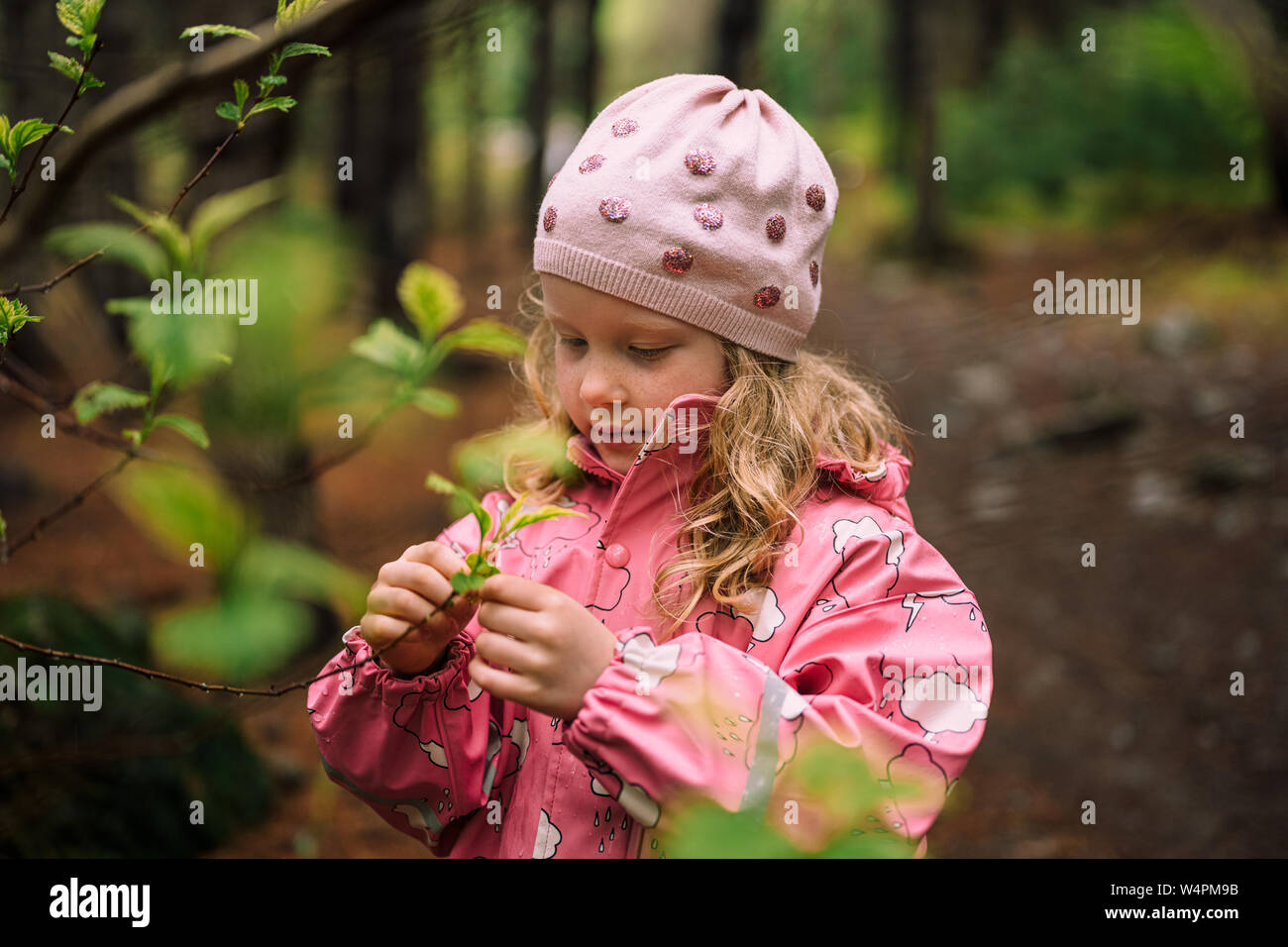 Adorable girl in pink jacket and cap attentively looking branch during